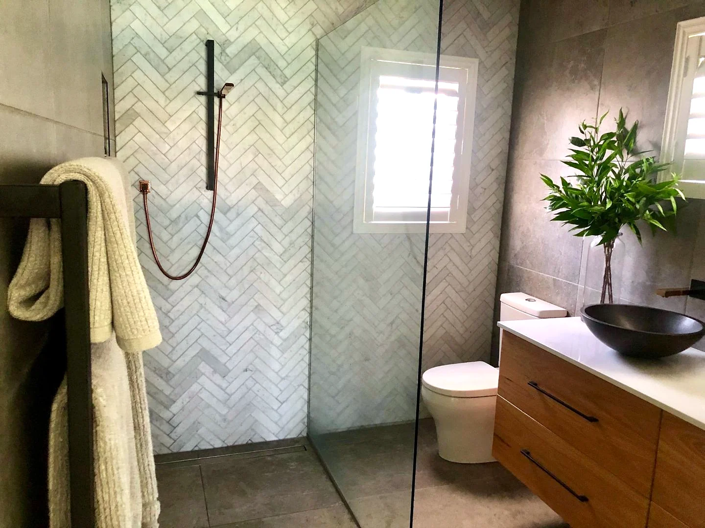 Bathroom with a marble herringbone shower feature wall, large-format grey floor tile, timber vanity with a dark stone vessel basin and plantation shutters