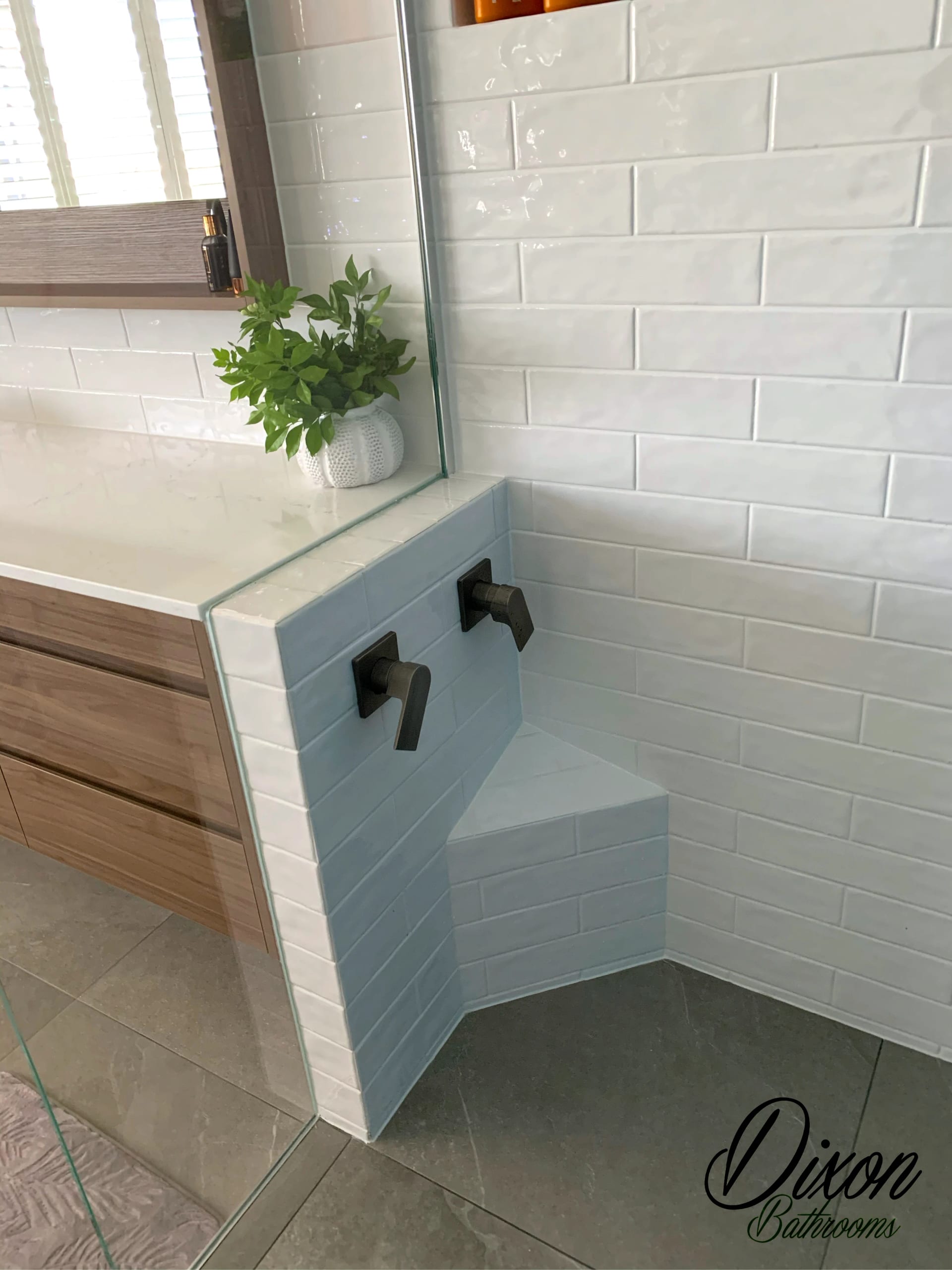 Walk-in shower with a tiled corner bench seat in pale blue subway tile, white gloss subway walls, matte black wall mixers and a frameless glass screen