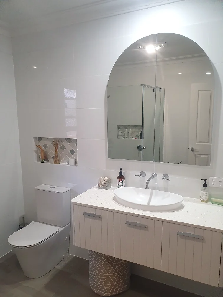White bathroom with an arched mirror, fish-scale mosaic tiled niche, vertical-groove vanity and an oval above-counter basin